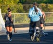 Women's field hockey team walking on track with goalie in blue jersey carrying hockey sticks.