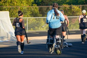 Women's field hockey team walking on track with goalie in blue jersey carrying hockey sticks.