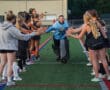Field hockey team cheers goalie running through teammates' tunnel on turf field.