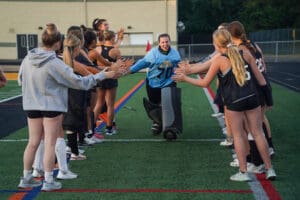Field hockey team cheers goalie running through teammates' tunnel on turf field.