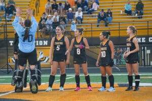 Field hockey team in black uniforms on the field, goalie jumping, crowd in the background.