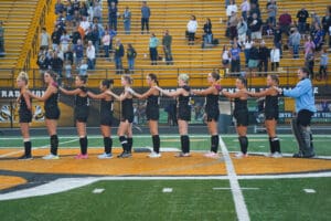 High school field hockey team stands in a line, preparing for a game, with spectators in the stadium.