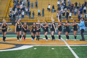 Girls' field hockey team celebrating on the field with cheering crowd in the background.