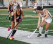Girls playing competitive field hockey match on a green turf field.