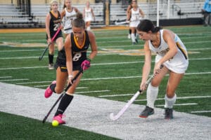 Girls playing competitive field hockey match on a green turf field.
