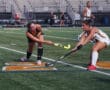 Two female field hockey players compete for the ball on a turf field during a match.