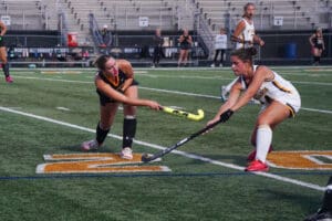 Two female field hockey players compete for the ball on a turf field during a match.