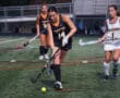 Field hockey player dribbles ball during match; opponent in pursuit on turf field.