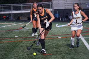 Field hockey player dribbles ball during match; opponent in pursuit on turf field.