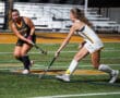 Two female field hockey players competing in a match at night, with focus on the ball on a green turf field.