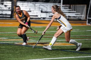 Two female field hockey players competing in a match at night, with focus on the ball on a green turf field.