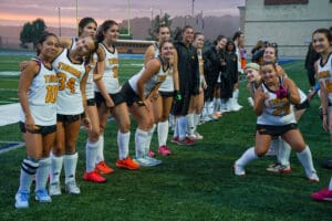 High school girls' field hockey team in uniform on the field, smiling and posing before a game at sunset.