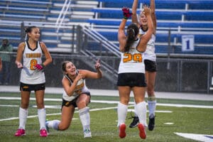 Field hockey team celebrates a victory with smiles and cheers on a rainy sports field.