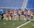 Girls' field hockey team joyfully running on the field during practice at a stadium with blue seats.