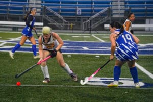 Field hockey match action with players competing for the ball on a grass field.