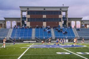 Field hockey game at Spartan Stadium during overcast weather with players in motion and spectators in the stands.