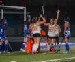 Field hockey players celebrate a goal at night; team in white and black with a vibrant orange goalie.