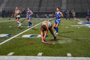 Field hockey players compete intensely in rain on a synthetic field, focusing on a red ball.