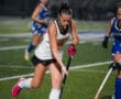 Girls playing intense field hockey match on turf, focusing on player in black and white jersey with the ball.