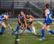 Four female field hockey players competing intensely at night on turf field.