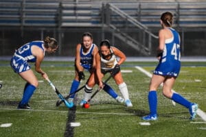 Four female field hockey players competing intensely at night on turf field.