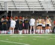 Field hockey team huddles on the sideline, preparing for a match on an outdoor field.