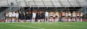Field hockey team huddles on the sideline, preparing for a match on an outdoor field.