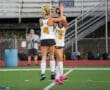 Two field hockey players in team uniforms high-fiving on the field.