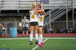 Two field hockey players in team uniforms high-fiving on the field.