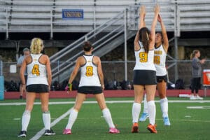 Four female field hockey players in warm-up on a field, numbered jerseys visible.