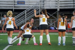 Field hockey team celebrating during a match, wearing white and black jerseys with numbers, on a green pitch.