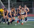 Field hockey players in action, competing for the ball on a green field, with goalie standing in the goal area.