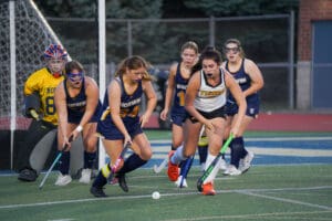 Field hockey players in action, competing for the ball on a green field, with goalie standing in the goal area.