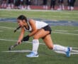 Field hockey player in action, wearing white and black uniform on a green turf field.
