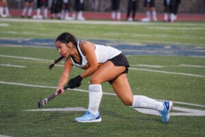 Field hockey player in action, wearing white and black uniform on a green turf field.