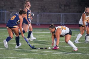 Women's field hockey match, two players battling for ball on turf field.