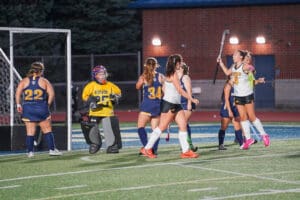 Field hockey players celebrating a goal on a lit outdoor field with goalie in action.