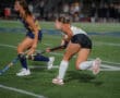 Two women playing a fast-paced field hockey game on a green turf field, focused on the ball.