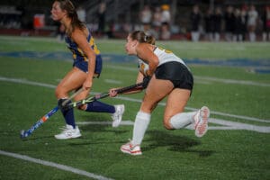 Two women playing a fast-paced field hockey game on a green turf field, focused on the ball.