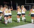 Field hockey team walking on turf, wearing numbered jerseys and holding sticks, preparing for a game at night.