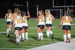 Field hockey team walking on turf, wearing numbered jerseys and holding sticks, preparing for a game at night.
