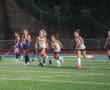 Girls playing field hockey at night, wearing blue and white uniforms, on a lit turf field.