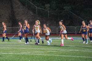 Girls playing field hockey at night, wearing blue and white uniforms, on a lit turf field.