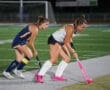 Two female field hockey players in action on a turf field at night, one in white and the other in blue.