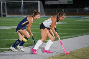 Two female field hockey players in action on a turf field at night, one in white and the other in blue.