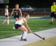 Field hockey player in white jersey prepares to hit ball during a match under bright stadium lights.