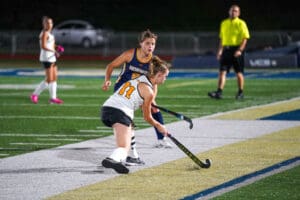 Field hockey players in action during a game, with referee in the background.