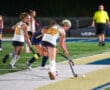 Field hockey players in action on artificial turf during a night game, with referee observing.