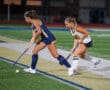 Two female field hockey players compete for the ball during a match on a turf field.
