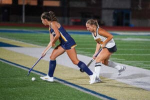 Two female field hockey players compete for the ball during a match on a turf field.